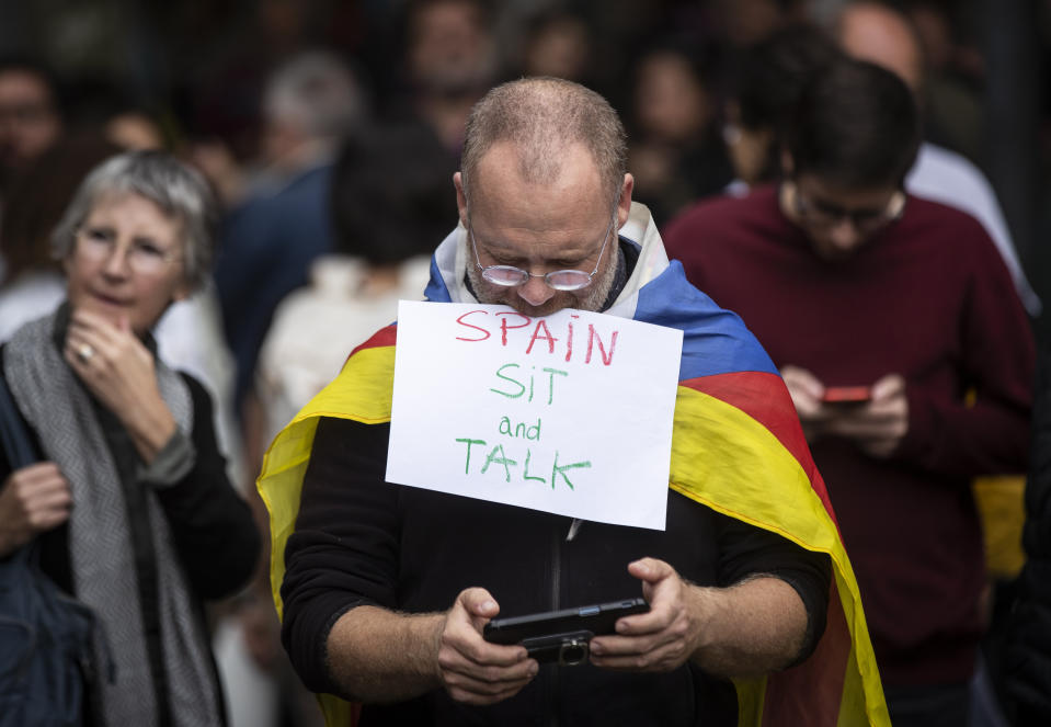 A Catalan pro-independence protester, wearing a Catalan independence flag, checks his phone after taking a picture as he and others demonstrate outside the building of the Government Delegation in the Autonomous Community of Catalonia, in downtown Barcelona, Spain Monday, Oct. 21, 2019. Spanish leader Pedro Sanchez is traveling to Barcelona Monday, the protest-struck capital of the northeastern Catalonia region, to visit with injured police officers and talks with officials in charge of security. (AP Photo/Ben Curtis)