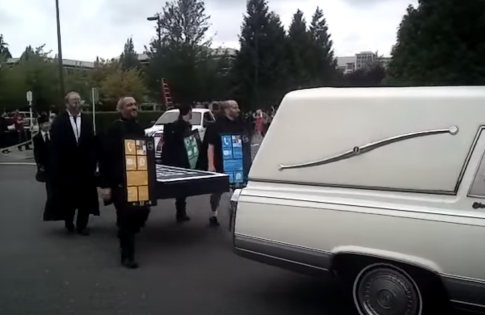 A hearse driving down a street followed by people dressed as Windows phones holding a big cardboard iPhone like a coffin
