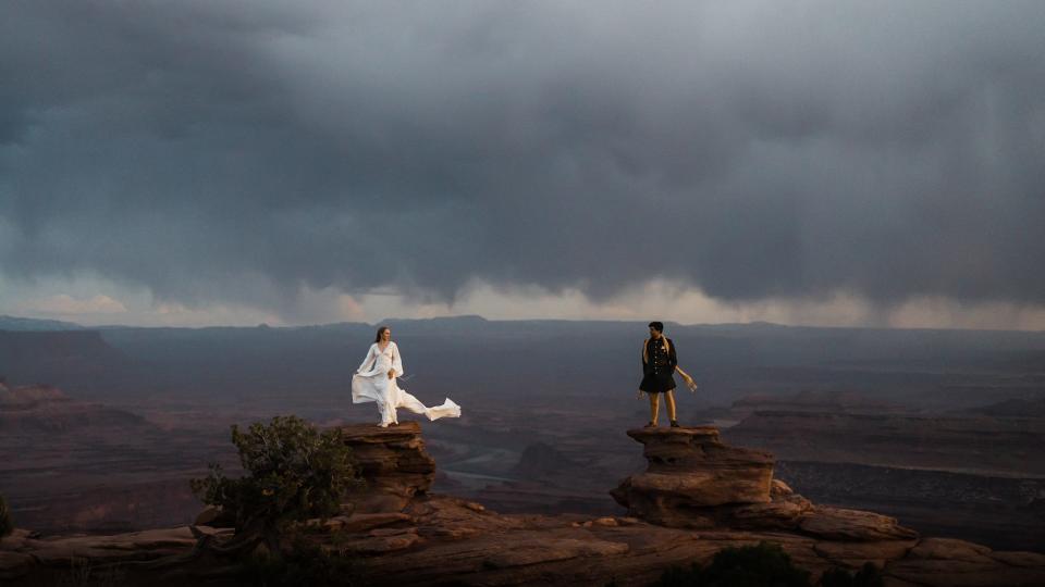 A bride and groom look at each other as they stand on two rocks in front of a cloudy landscape.