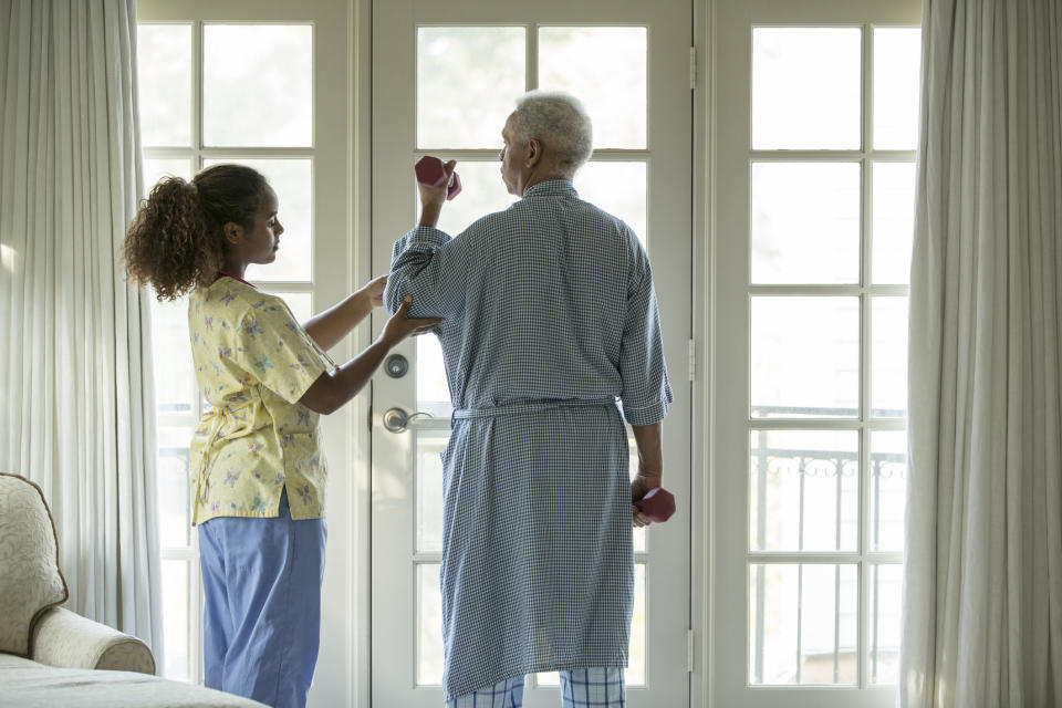 A woman helps an older man in a robe use hand weights by a window