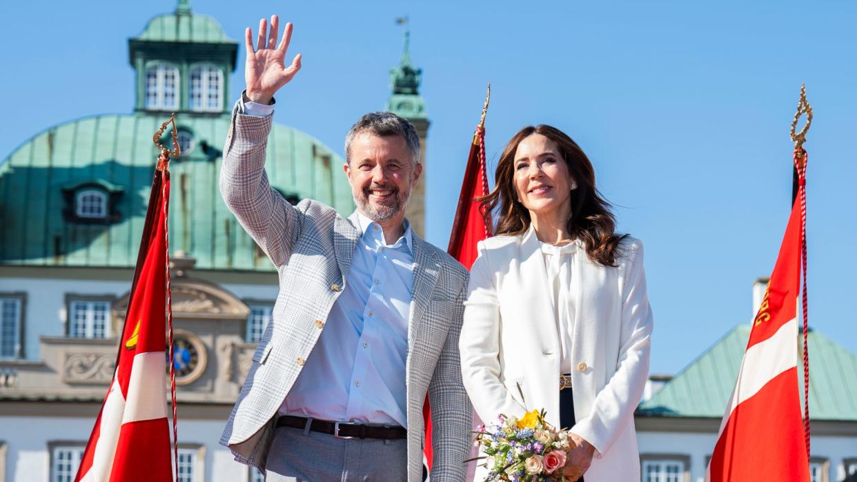 King Frederik waving and standing with wife Queen Mary who holds a bunch of flowers