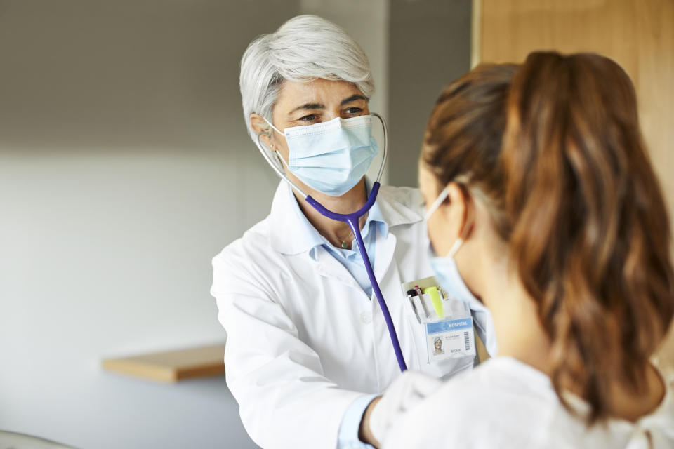 A healthcare worker with short, gray hair uses a stethoscope to check the heart rate of a patient wearing a ponytail, both of them wearing surgical masks