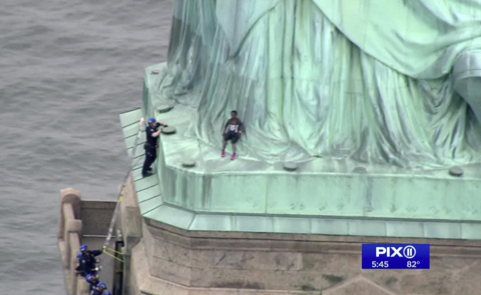 In this image made from video by PIX11, a person, center, leans against the robes of the Statue of Liberty on Liberty Island in New York, as one of the police officers climbed up on a ladder to stand on a ledge nearby talking the climber into descending on Wednesday, July 4, 2018. Shortly before the climber scaled the statue’s base, forcing its evacuation, several people who hung a banner from the Statue of Liberty’s pedestal calling for abolishing the federal government’s chief immigration enforcement agency were arrested. (PIX11 via AP)