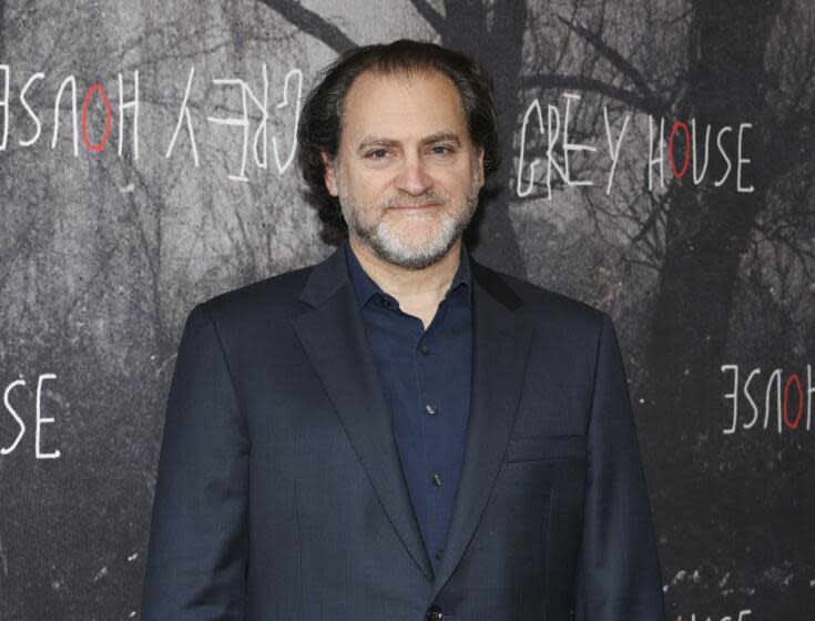 A man with short dark hair, white facial hair in a dark suit standing against a dark gray backdrop