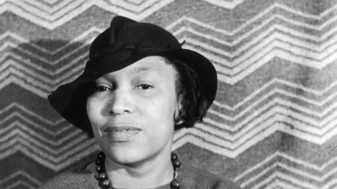 zora neale hurston wearing a sweater and hat and sitting for a photo in front of a striped backdrop