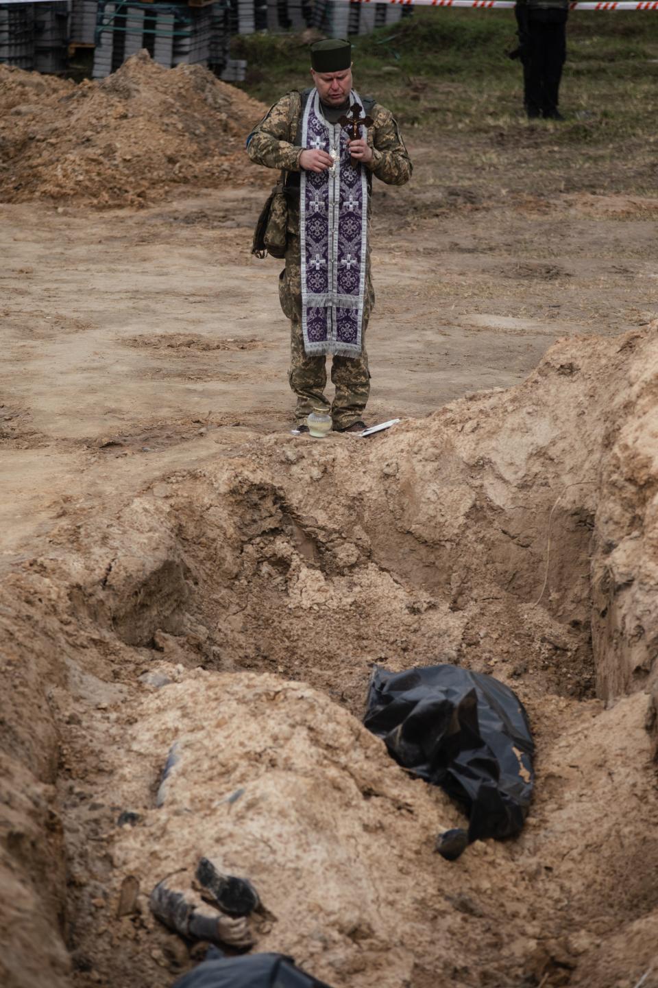 An religious figure with a long robe appears to bless a ditch where bodies, contained in bags, lie below.