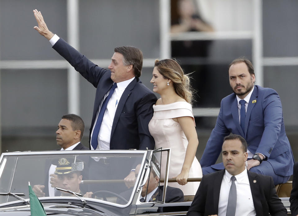 FILE - In this Jan 1, 2019 file photo, flanked by first lady Michelle Bolsonaro, center, and his son Carlos Bolsonaro, right, Brazil's President Jair Bolsonaro waves as he rides in an open car after his swearing-in ceremony, in Brasilia, Brazil. Carlos Bolsonaro's tweet on Monday, Sept. 10, has caused an uproar saying democracy is slowing development in Latin America's biggest country. (AP Photo/Andre Penner, File)