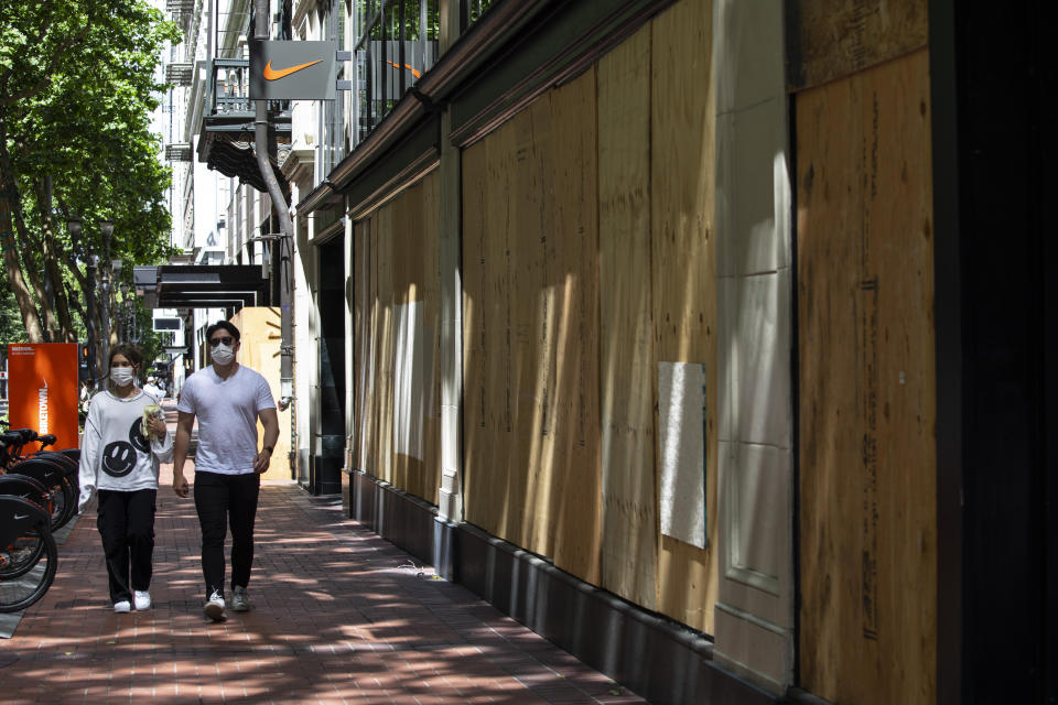 A couple walks past the the Nike store on Saturday, June 5, 2021, which is protected by wooden panels because of on-going protests in downtown Portland, Ore. City officials insist Portland is resilient as they launch a revitalization plan — in the form of citywide cleanups of protest damage, aggressive encampment removals, increased homeless services and police reform — to repair its reputation. (AP Photo/Paula Bronstein)