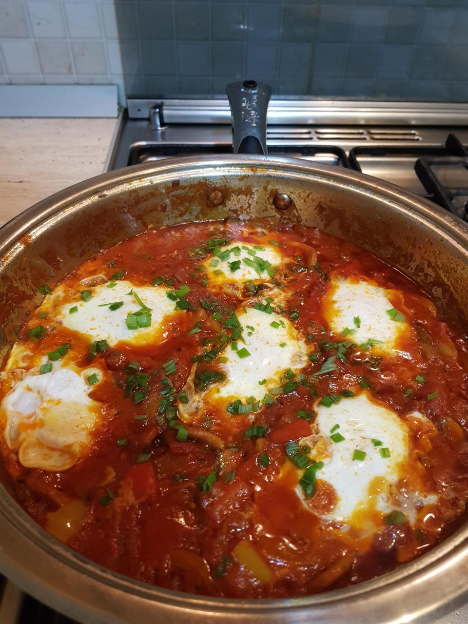 A pan of shakshuka, a dish with poached eggs in a tomato and pepper sauce, cooked with spices and garnished with chopped green herbs