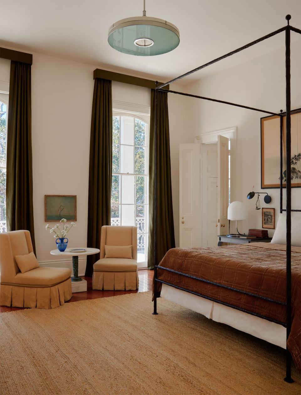 a four poster wrought iron bed has a chestnut brown cover, two armless chairs upholstered in linen with a round marble top side table between them, windows with deep green linen curtains, braided jute rug