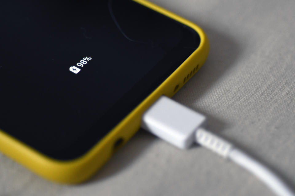 A cellphone with a yellow case, its battery charger plugged in, noting that the battery is 98% charged. 