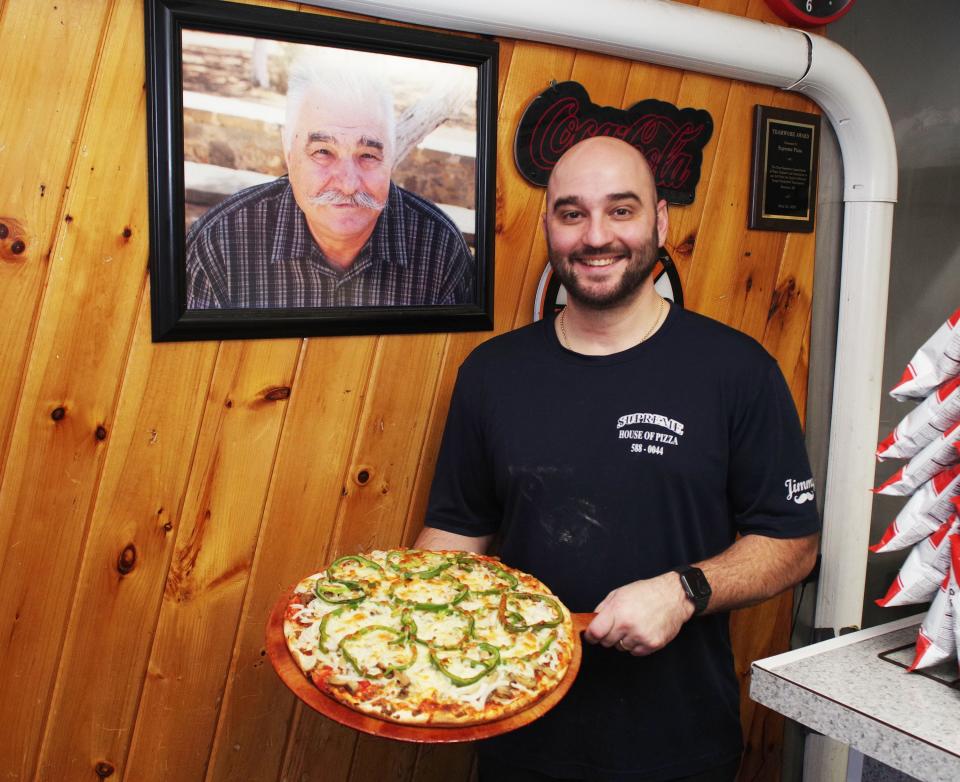 Supreme House of Pizza owner Manny Lambrakis, on Tuesday, Nov. 14, 2023, holding a pizza with cheese, green peppers and other ingrediants, stands next to a picture of his dad, Jimmy Lambrakis, who founded the restaurant in 1972 and worked in it right up to his passing in 2016.