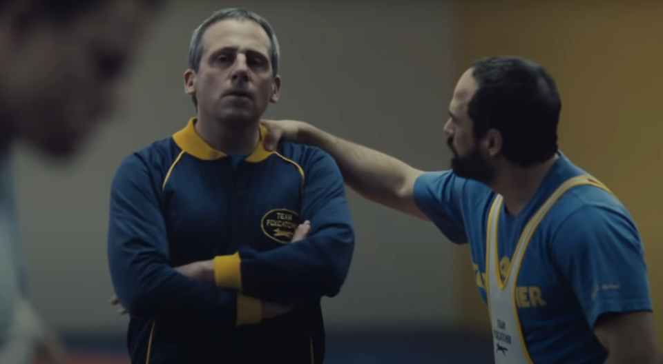 Steve Carell and Mark Ruffalo in workout clothes are standing in an indoor sports setting. The man is touching Carell's shoulder, and Carell looks contemplative