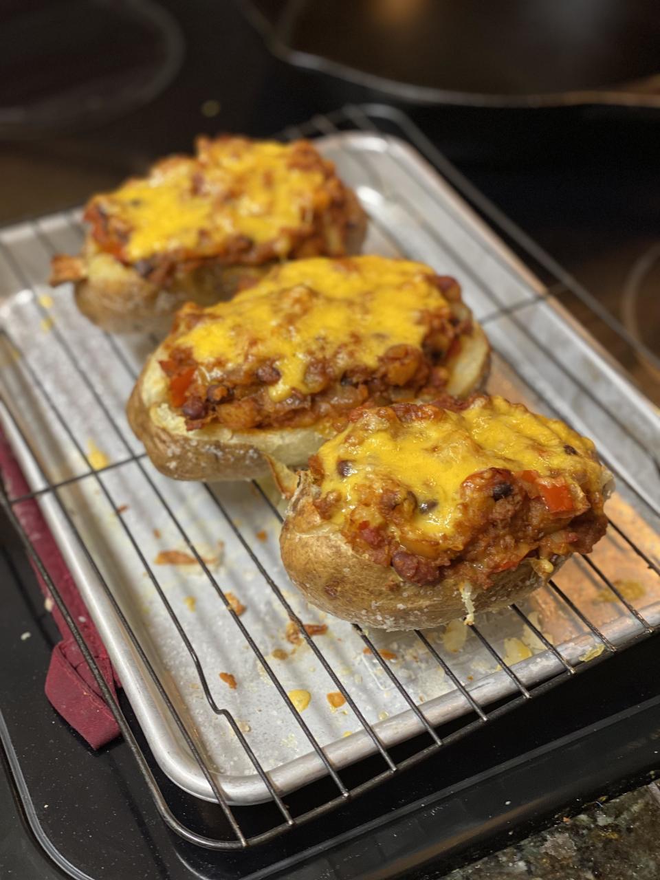 Three baked potatoes topped with melted cheese and a savory meat mixture, placed on a metal rack over a baking sheet