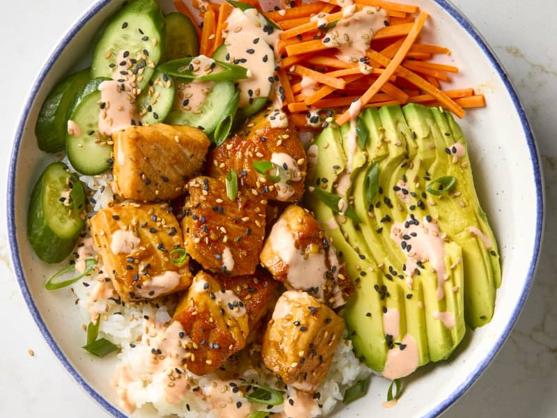 overhead shot of a salmon bowl with cucumbers, carrots and avocados, topped with black and white sesame seeds and spicy mayo