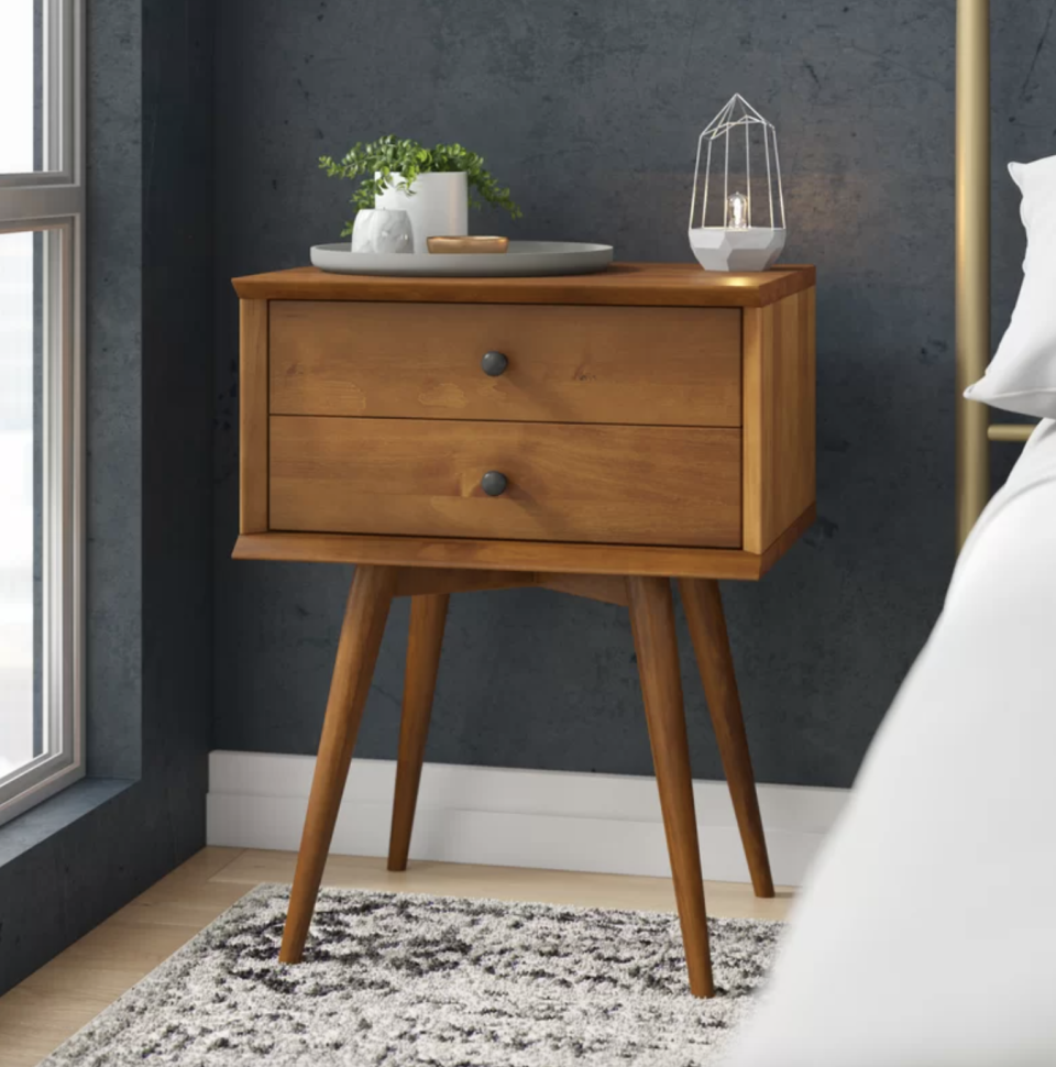 Grady 2 - Drawer Solid Wood Nightstand against grey wall and white and black carpet (Photo via Wayfair)