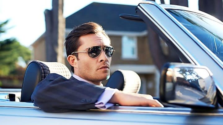 A man sitting in seat of a car with his arm out the window, top down with sunglasses on in a scene from White Gold.