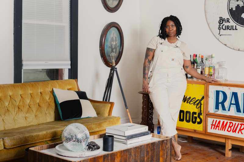 Barefoot dweller in overalls standing in white living room with tripod lamp, oval wood framed photos, wood coffee table, gold velvet couch, and sideboard decorated with signs.