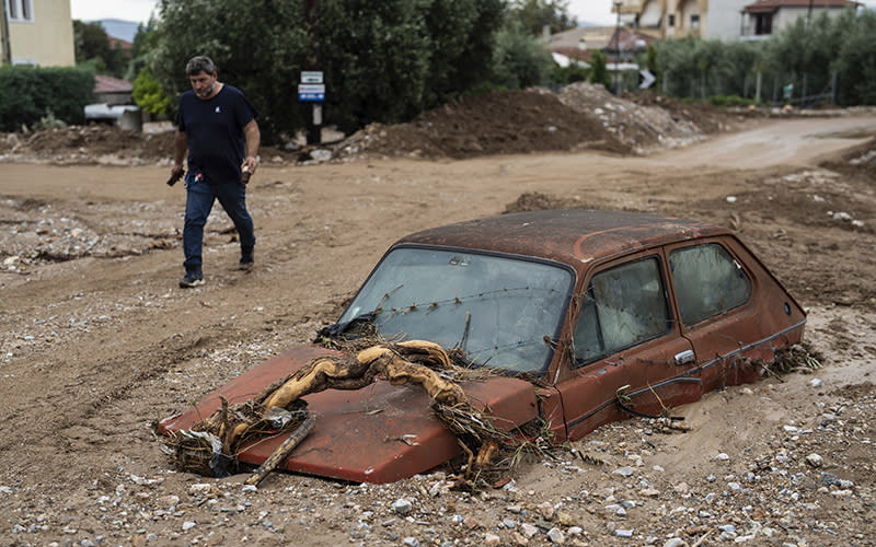 A red car is partially sunk into the ground after a flood. The ground around it is brown and muddy, with rocks and branches around. A man walks on a dirt path beside the vehicle.