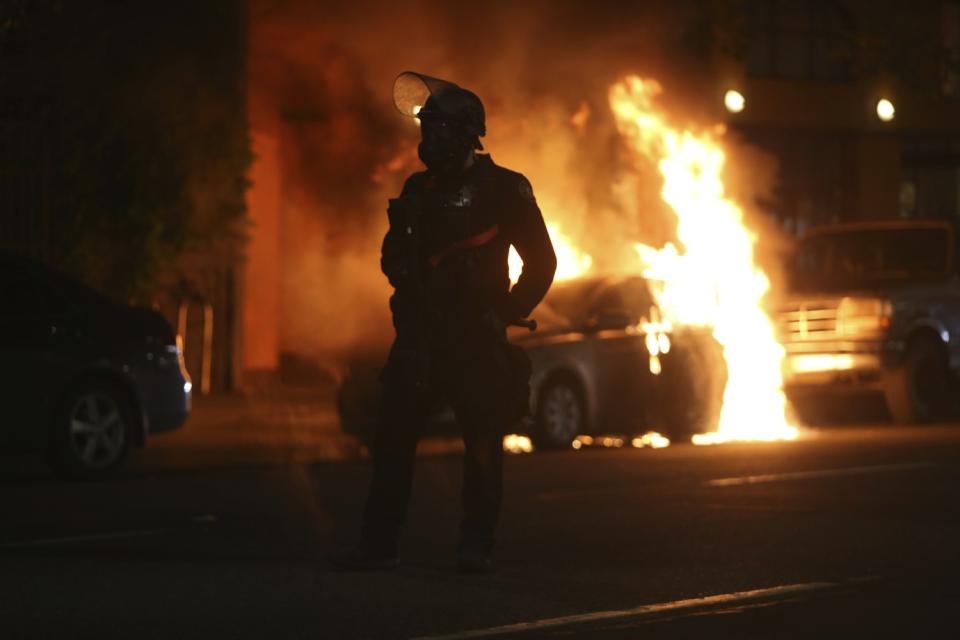 A police officer is silhouetted near a burning car.