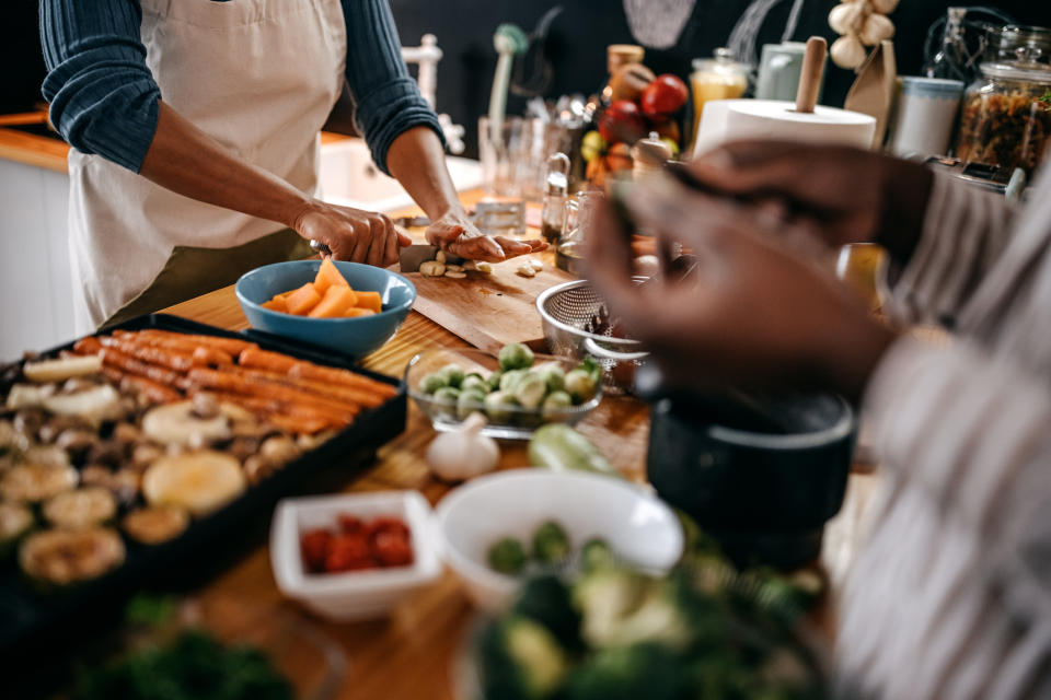 Two people prepare various vegetables in a kitchen, including carrots, zucchini, Brussels sprouts, and tomatoes, with bowls and cutting boards on a wooden countertop