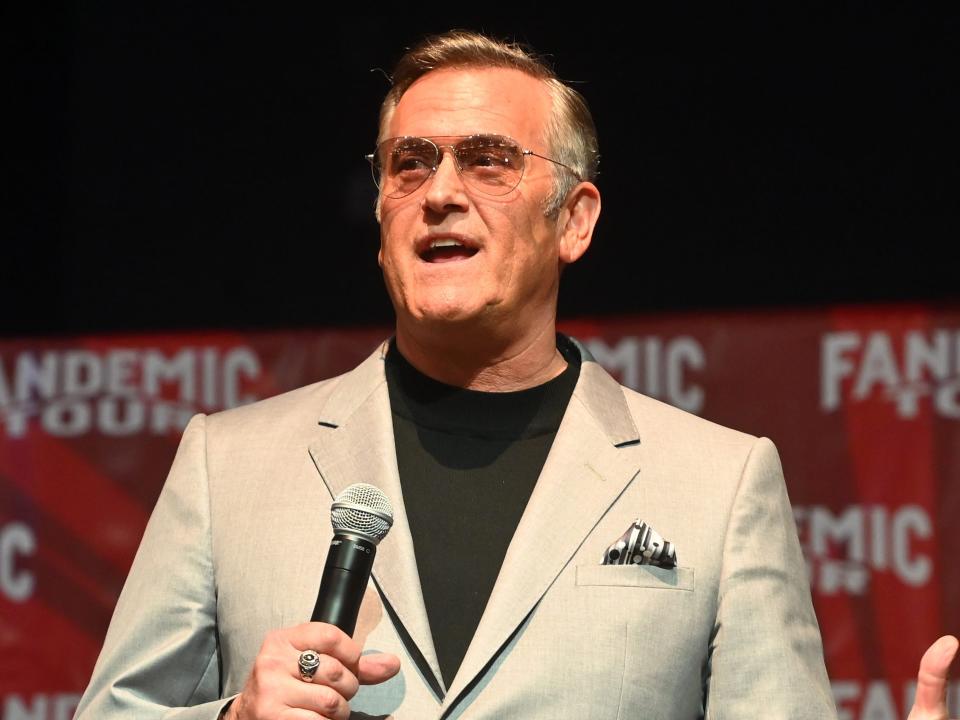 Bruce Campbell holds a microphone on stage.