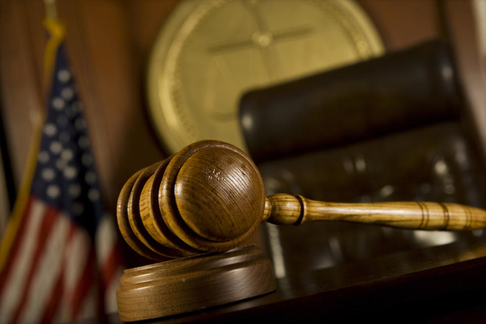 A judge's gavel up close, with the judge's chair, the American flag, and the courtroom seal in the background.