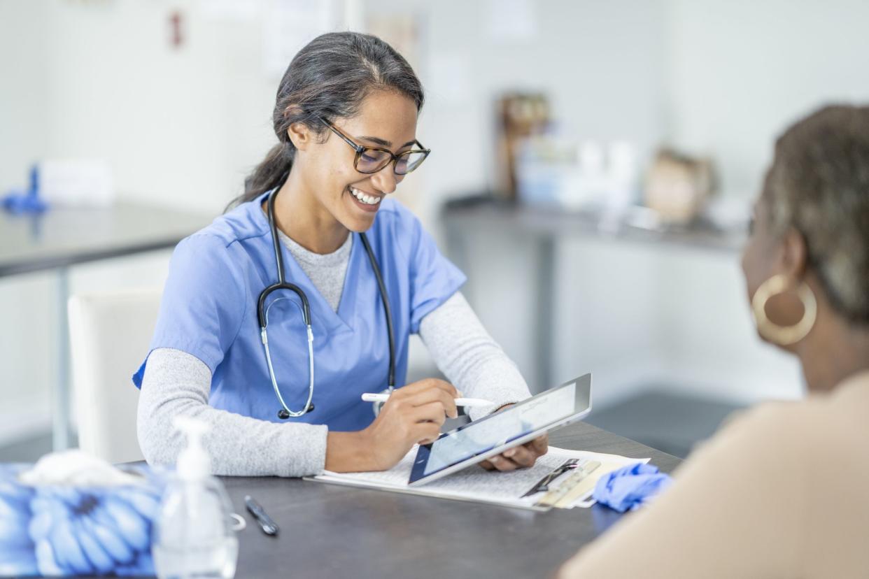 Female nurse practitioner smiling while on tablet with female patient during a medical appointment while at a desk with a blurred background of an exam room