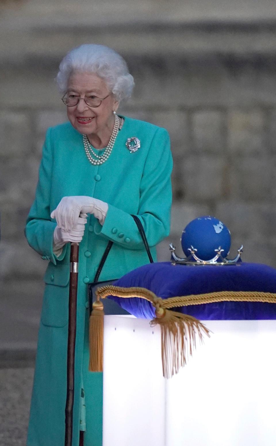 The Queen standing and smiling in a minty jade green long coat with buttons and pockets. She has on pearls, a round brooch, a black handbag, and is resting on a walking cane.