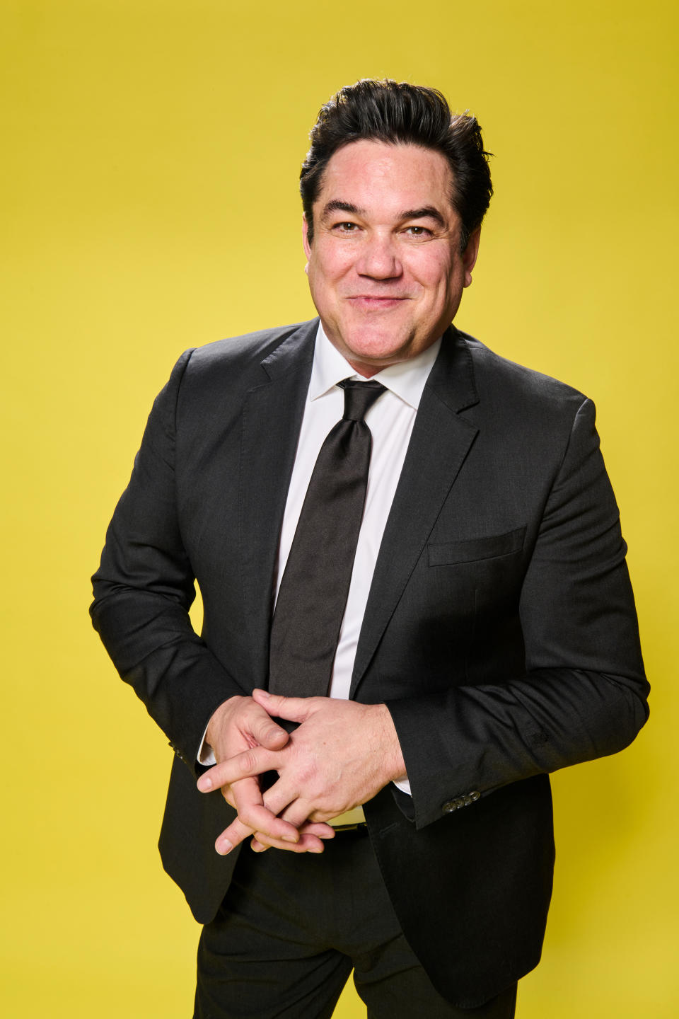 Man in suit with hands clasped, smiling against a yellow background
