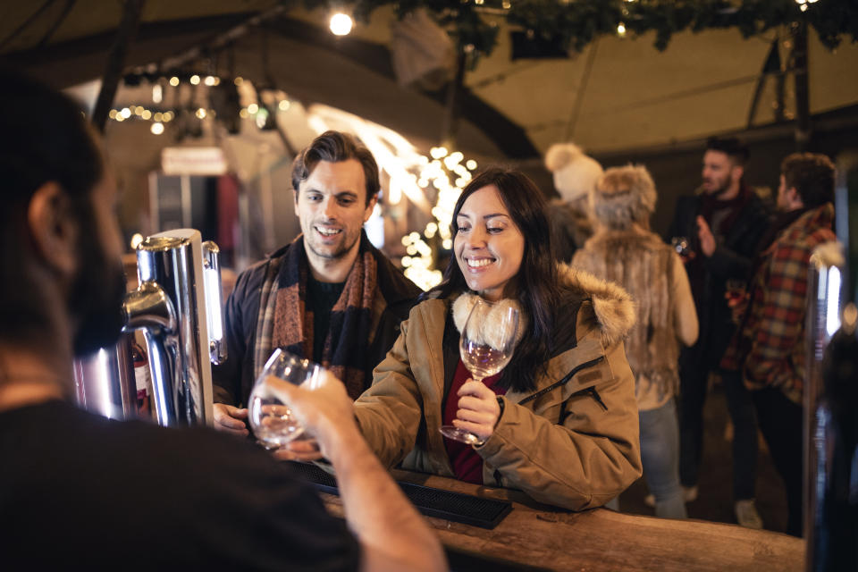 Rear view of a bar man handing a wine glass over to a mid adult woman. The woman is standing with a mid adult man. In the background there is a group of friends standing and talking.