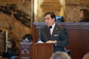 Rep. Nicholas Muscarello standing at a podium with a microphone in a dark grey suit.