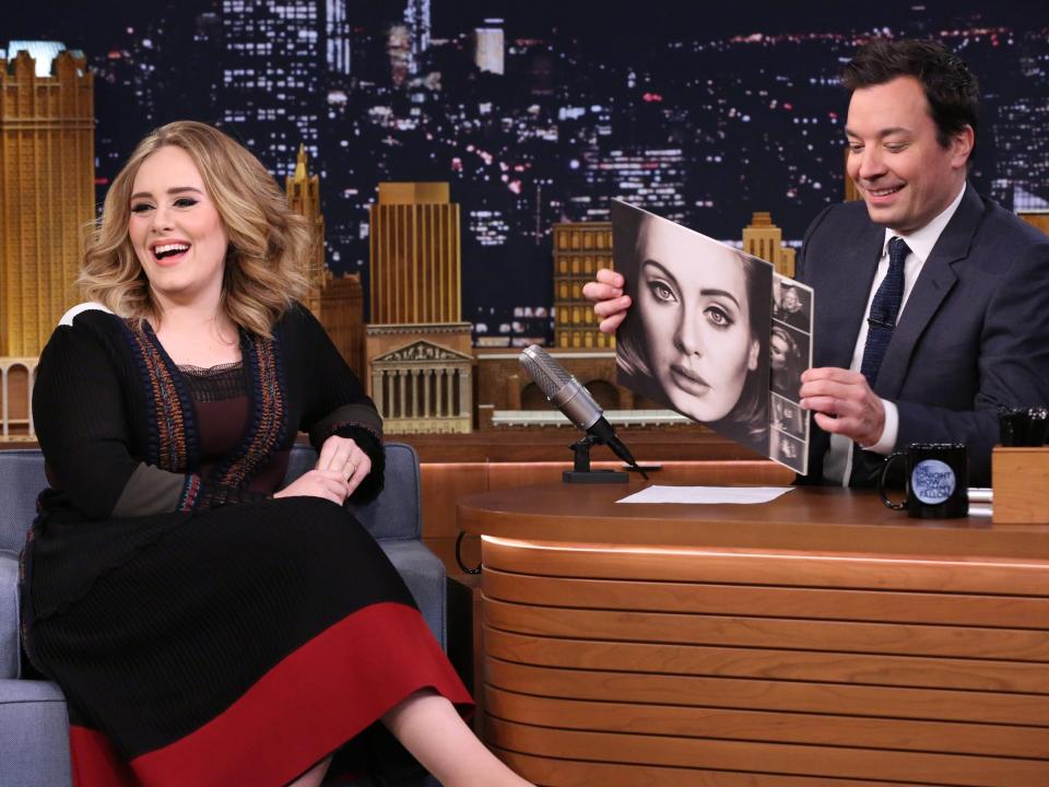 Adele smiling and laughing will sitting on a chair next to Jimmy Fallon behind his desk holding her album vinyl with her face on the cover.