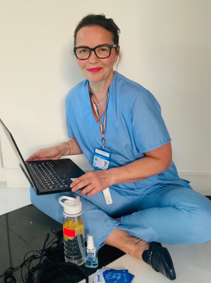 Vera Ora, an NHS psychiatrist, sits on the floor wearing blue hospital scrubs with a laptop on her knee
