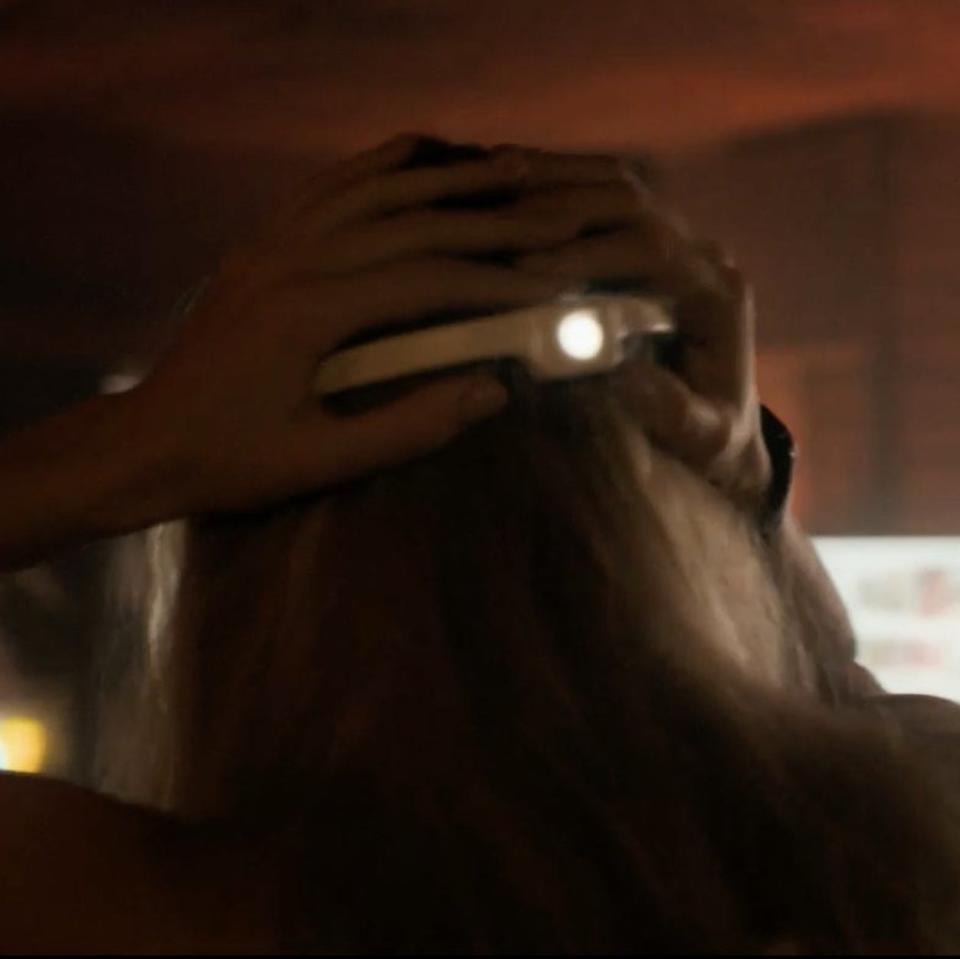 Sasha Luss wearing a white headband with a lashlight in darkness, facing away, with indistinct background suggesting an intense scene