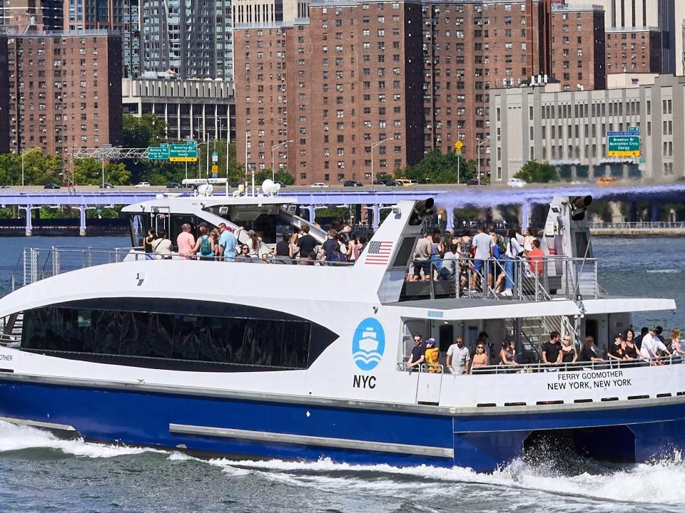 A crowded NYC Ferry travels on the East River, Behind is the Manhattan Bridge and the skyscrapers of Lower Manhattan on a clear summer day.