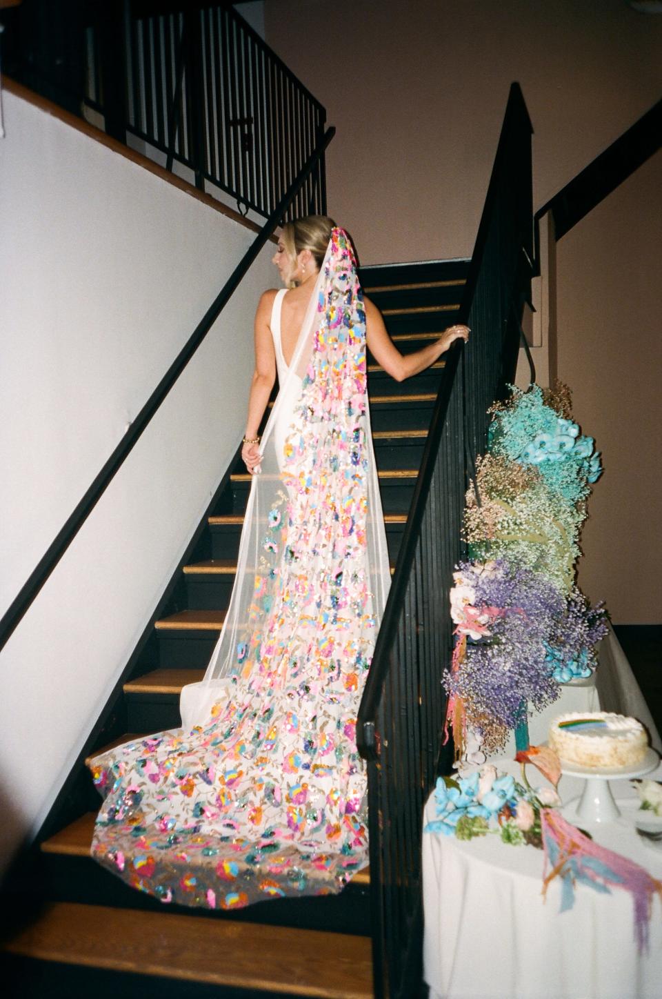 A bride in a white dress and rainbow veil walks up a staircase.