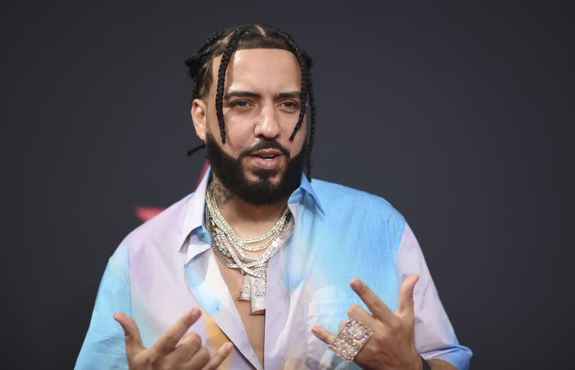 A man with braids, a beard and gold chains over a multicolored shirt makes gestures with both hands