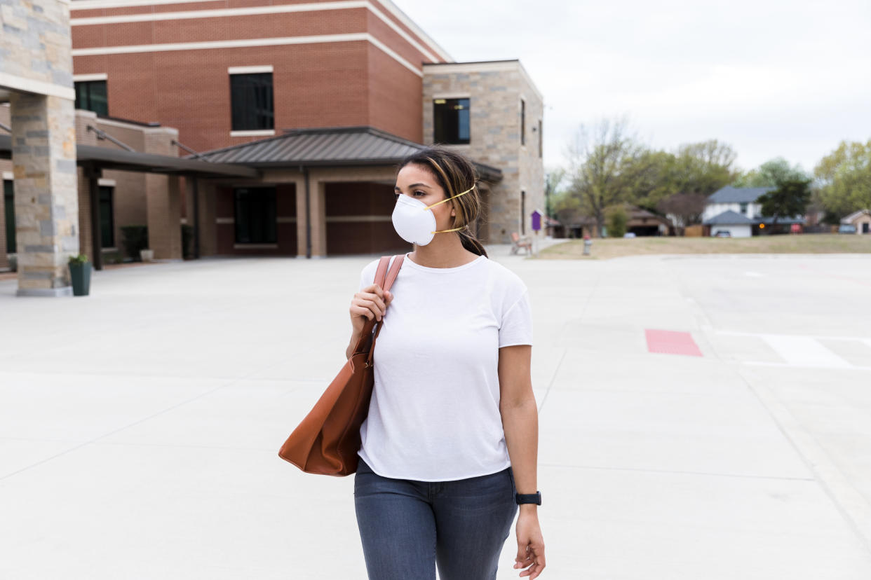 A mid adult female teacher walks by her empty school during the epidemic.  She is wearing a mask for safety.