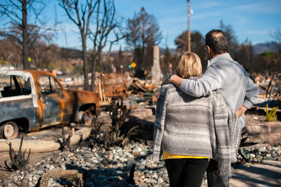woman and man with arms around each other looking at burned out house site