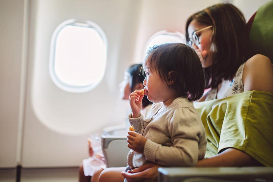 Little toddler girl having snacks joyfully while sitting on her mom’s lap on the airplane