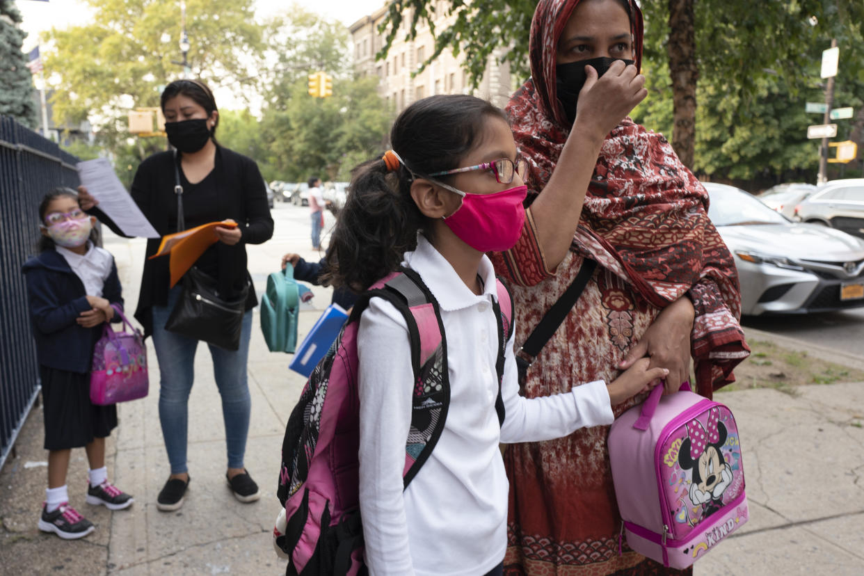 Children with backpacks and lunch containers stand with their parents on a sidewalk, all wearing face masks.