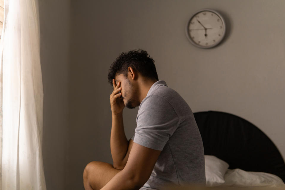 A person sits on a bed, covering their face with one hand, looking contemplative. A clock is on the wall behind them