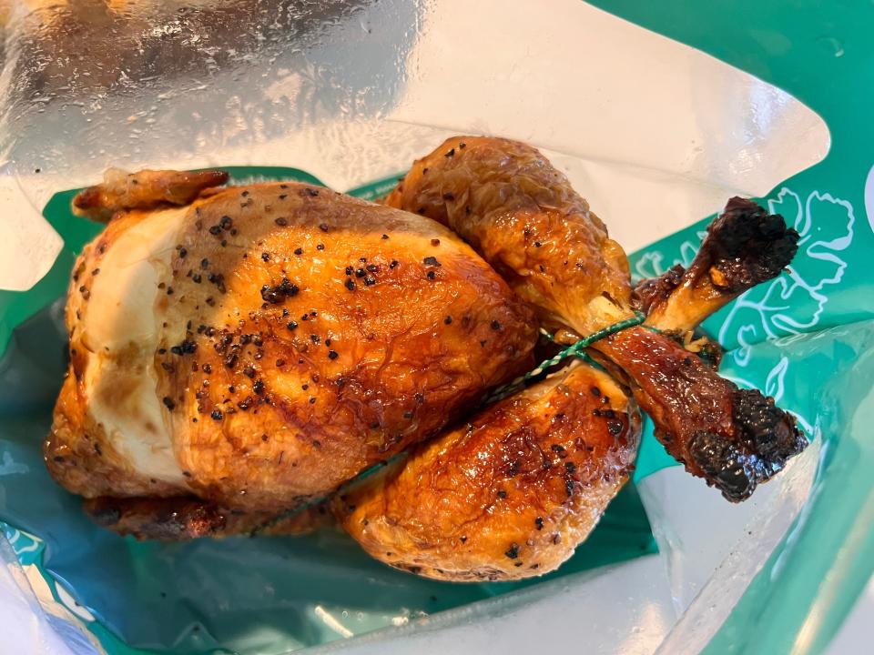 A rotisserie chicken sits in a bag with clear and green plastic. The legs of the chicken are tied together