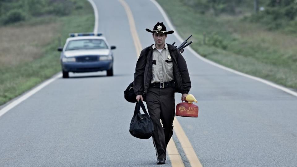 Andrew Lincoln as Rick Grimes wearing Rick's Hat and walking down an empty street with a police car in the background