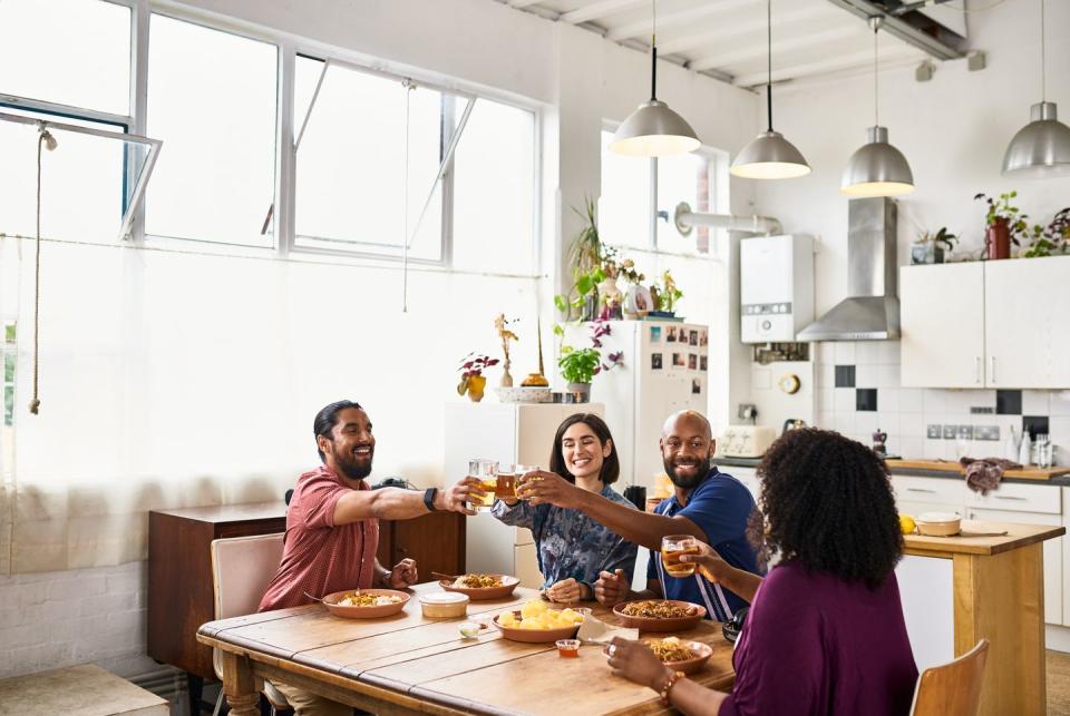 mid adult men and women sitting at kitchen table with takeaway meal, raising glasses for a toast, celebration, reunion, new normal, staying in