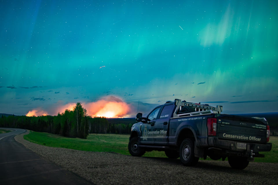 The wildfire forcing thousands to evacuate from a northeast British Columbia town has doubled again in size as the blaze grows merely a few kilometres west of city limits. The Aurora Borealis shines overhead of a B.C. Conservation Officer Service vehicle near the junction of highways 97 and 77, as a wildfire burns in the background near Fort Nelson, B.C., in a Saturday, May 11, 2024, handout photo. THE CANADIAN PRESS/HO-Ministry of Water, Land and Resource Stewardship, *MANDATORY CREDIT*