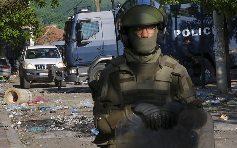 A KFOR soldier stands in the foreground; behind him are debris and Kosovo police officers guarding a municipal building after clashes