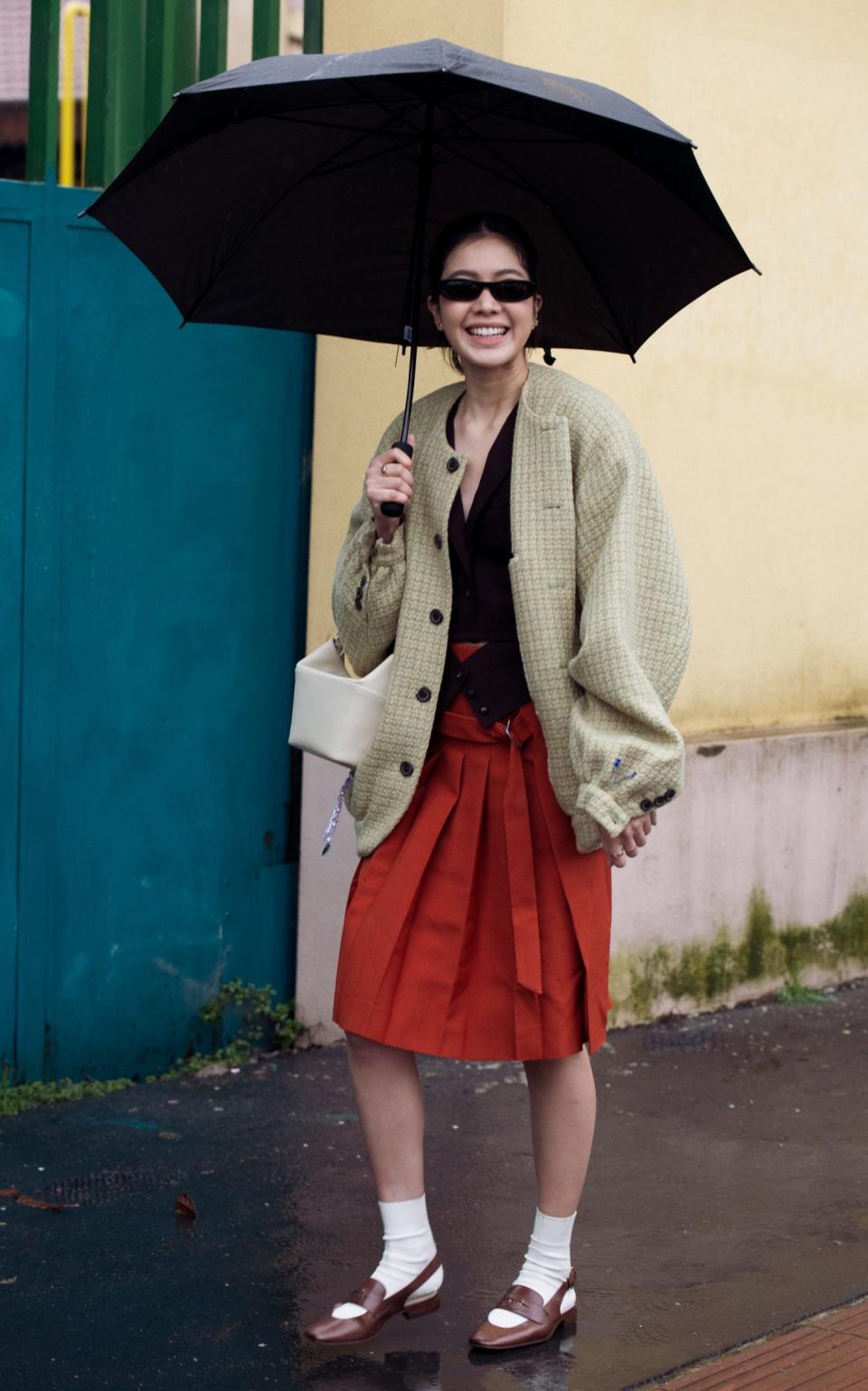 A guest at Milan Fashion Week wearing a red skirt, black vest, khaki jacket, white socks, brown shoes and beige bag