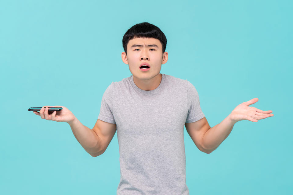 Unhappy young Asian man shrugging shoulder having trouble with his smartphone isolated on light blue studio background